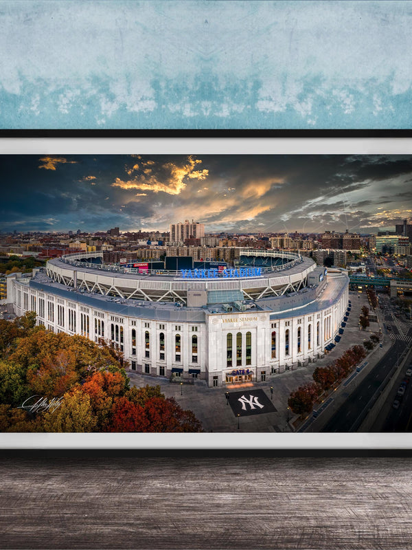 The image is a framed photograph of the New York Yankees stadium, which is a large, circular stadium located in the city of New York. The photograph captures the stadium's distinctive architecture and surrounding cityscape, with a dramatic sky in the background.