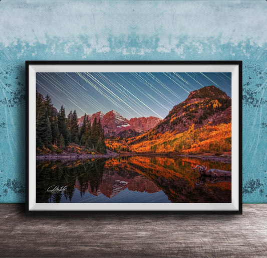 The image is a framed photograph depicting a serene mountain landscape with a lake reflecting the vibrant colors of the autumn foliage. The sky is filled with long exposure star trails, creating a stunning visual effect that adds depth and dimension to the scene.