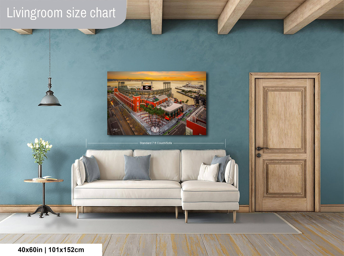 a living room with a large wall-mounted painting depicting a cityscape with a ferris wheel and buildings. The painting is framed and hangs above a white sofa with a wooden coffee table in front of it. The room has a wooden door and a wooden beam ceiling, creating a cozy and inviting atmosphere.