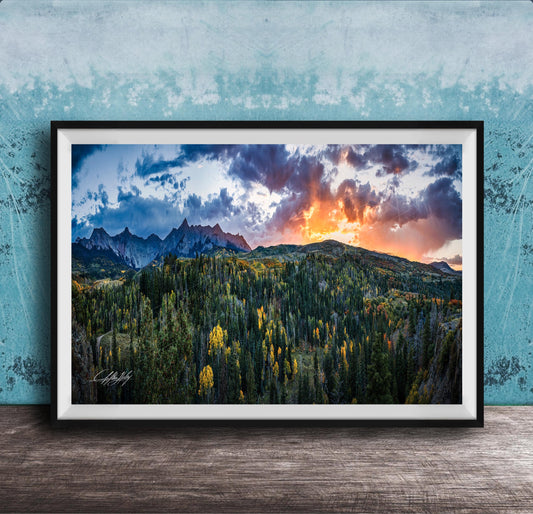 The image is a framed landscape photograph depicting a mountainous, forested area with a dramatic sky at sunset.