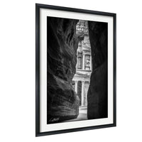 A black and white photograph of a narrow passage between two large rock formations, with the entrance to a building visible through the opening.