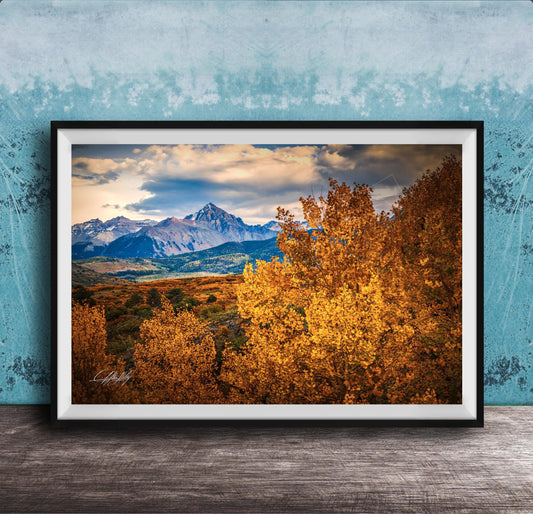 The image is a framed photograph depicting a scenic mountain landscape with a mix of autumn-colored trees and snow-capped peaks in the background.