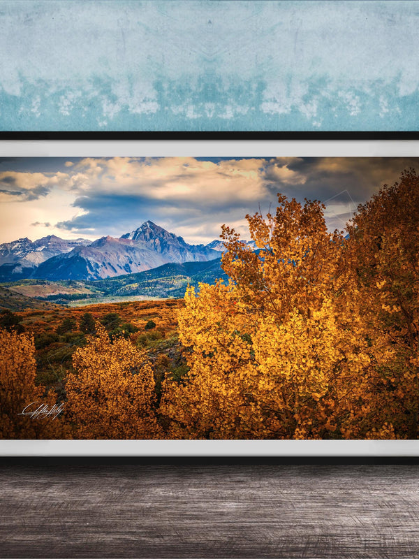 The image is a framed photograph depicting a scenic mountain landscape with a mix of autumn-colored trees and snow-capped peaks in the background.