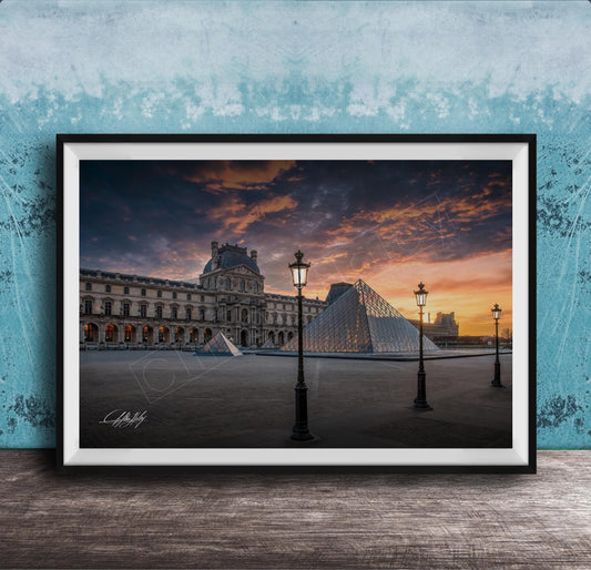 A framed photograph of the Louvre Museum in Paris, France, with the iconic glass pyramid structure of the Louvre Museum prominently featured against a dramatic sky at sunset.