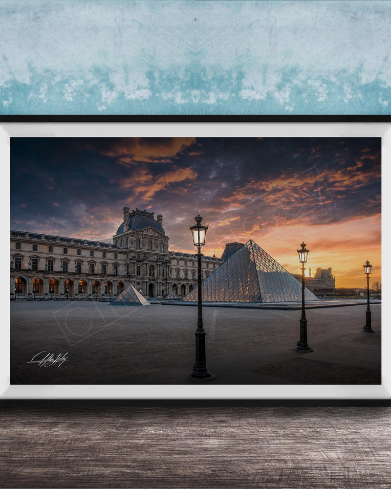 A framed photograph of the Louvre Museum in Paris, France, with the iconic glass pyramid structure of the Louvre Museum prominently featured against a dramatic sky at sunset.