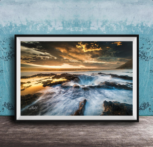 The image is a framed photograph of a rocky coastline with crashing waves, set against a dramatic sky with clouds and a sunset.
