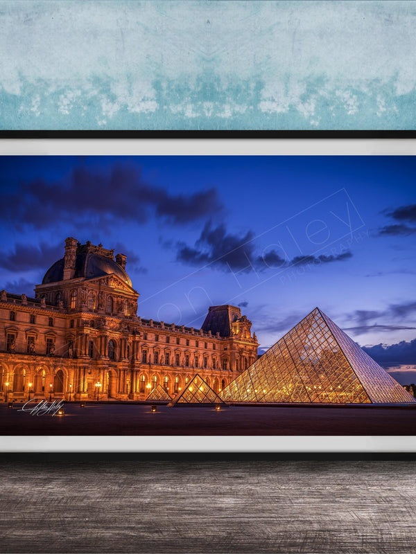 the Louvre Museum in Paris, France, with its iconic glass pyramid structure illuminated at night against a dark blue sky.