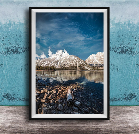 A framed photograph of a serene mountain landscape with a calm lake in the foreground, surrounded by snow-capped peaks and a clear blue sky.