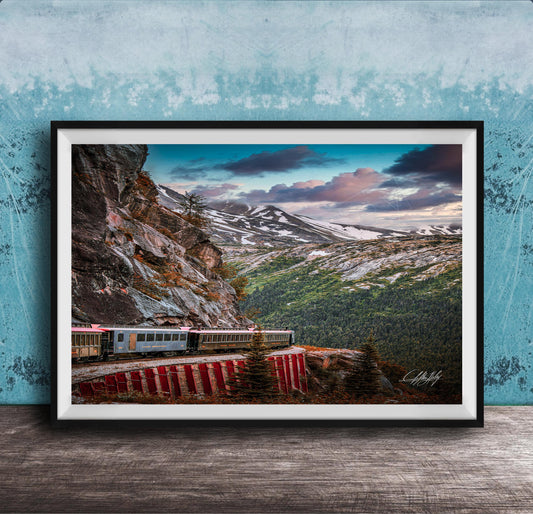A framed photograph depicting a train traveling along a winding track through a mountainous, forested landscape with snow-capped peaks in the background.