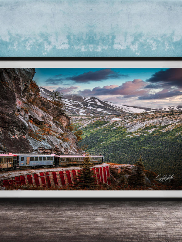 A framed photograph depicting a train traveling along a winding track through a mountainous, forested landscape with snow-capped peaks in the background.