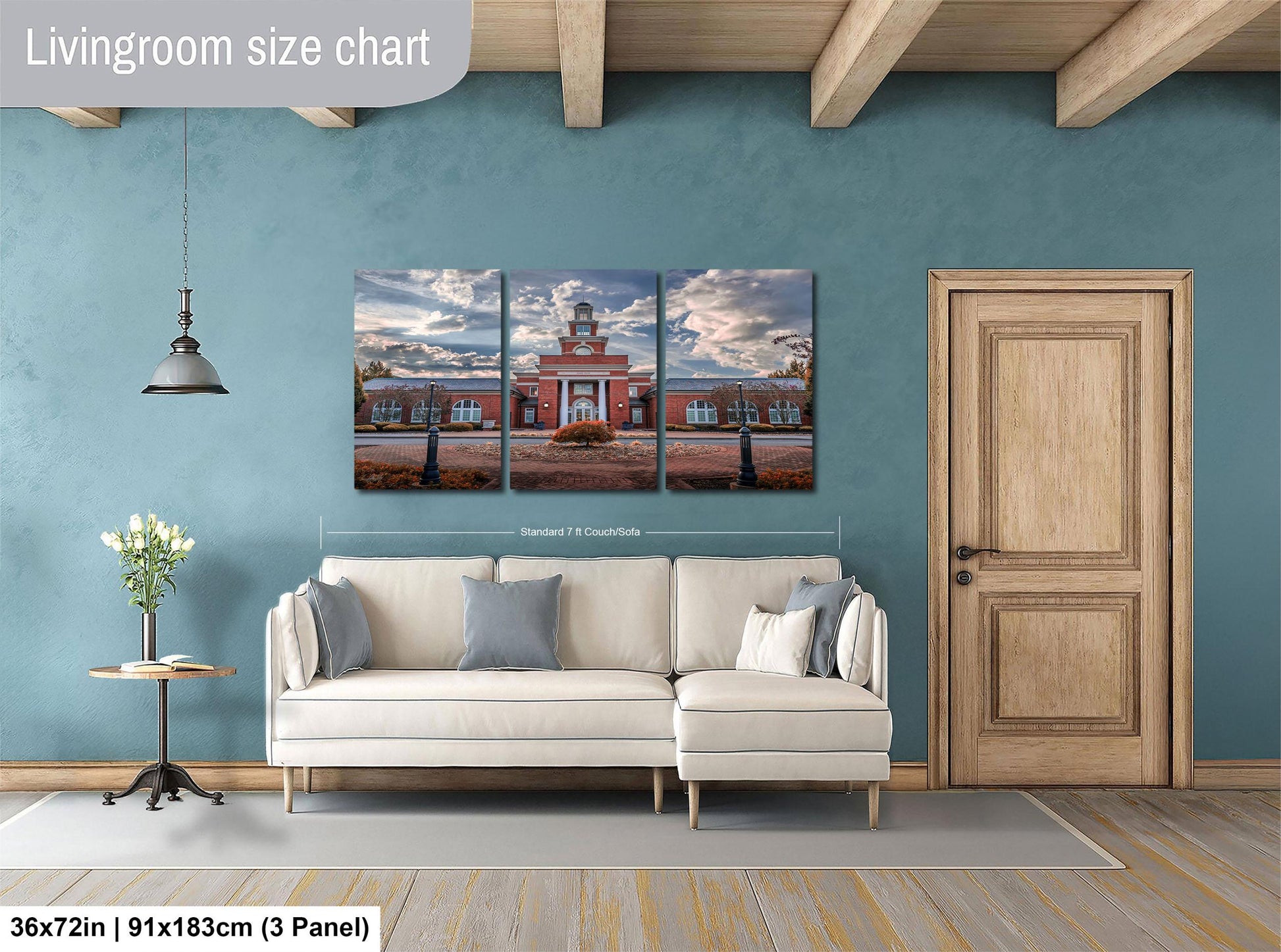The image is a living room with a large wall art depicting a red brick building with a clock tower. The wall art is divided into three panels, each showing different angles of the building. The living room has a white sofa and a wooden door, and there is a potted plant and a vase on a table. The wall art is approximately 36 inches wide and 9 inches tall.