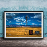A framed photograph depicting a windmill standing in a field with a blue sky and clouds in the background.