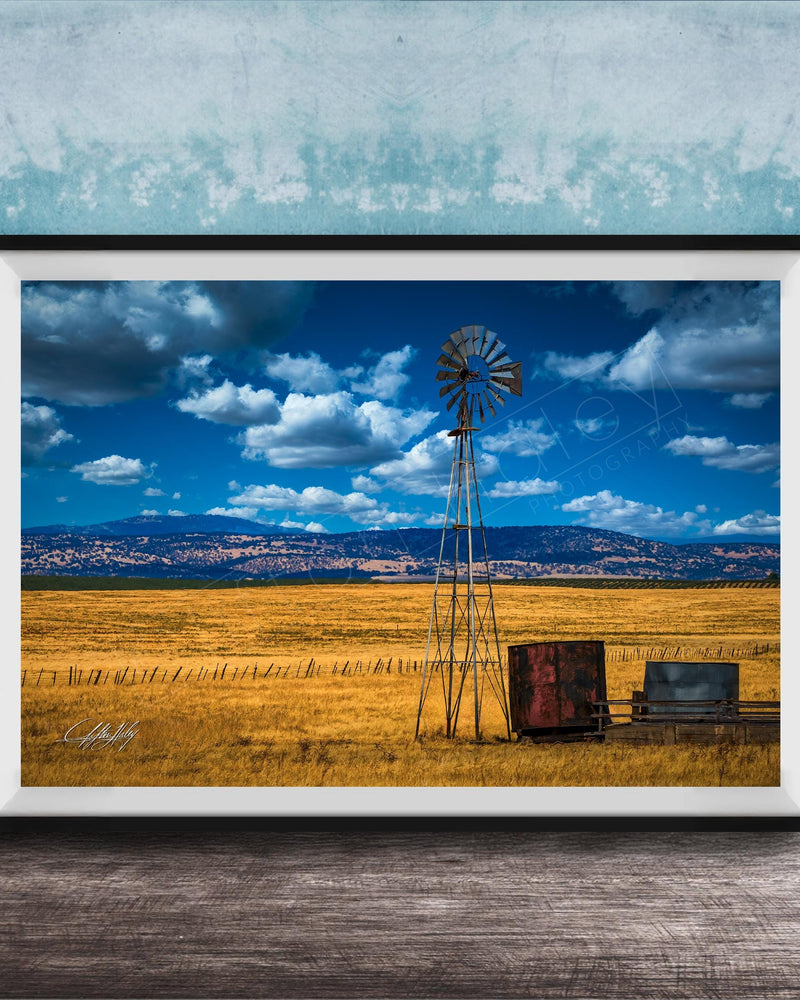 A framed photograph depicting a windmill standing in a field with a blue sky and clouds in the background.
