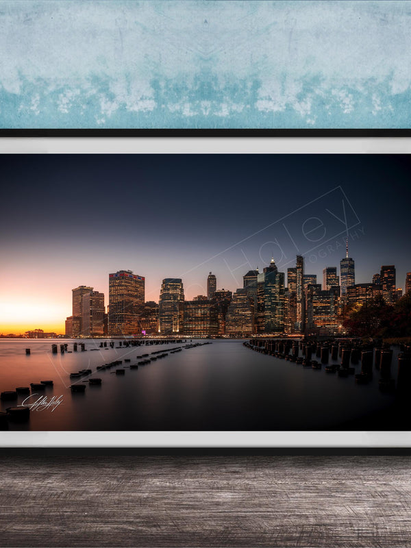 A framed photograph of the skyline of New York City at sunset, with the city's skyscrapers and landmarks illuminated against a dark blue sky.