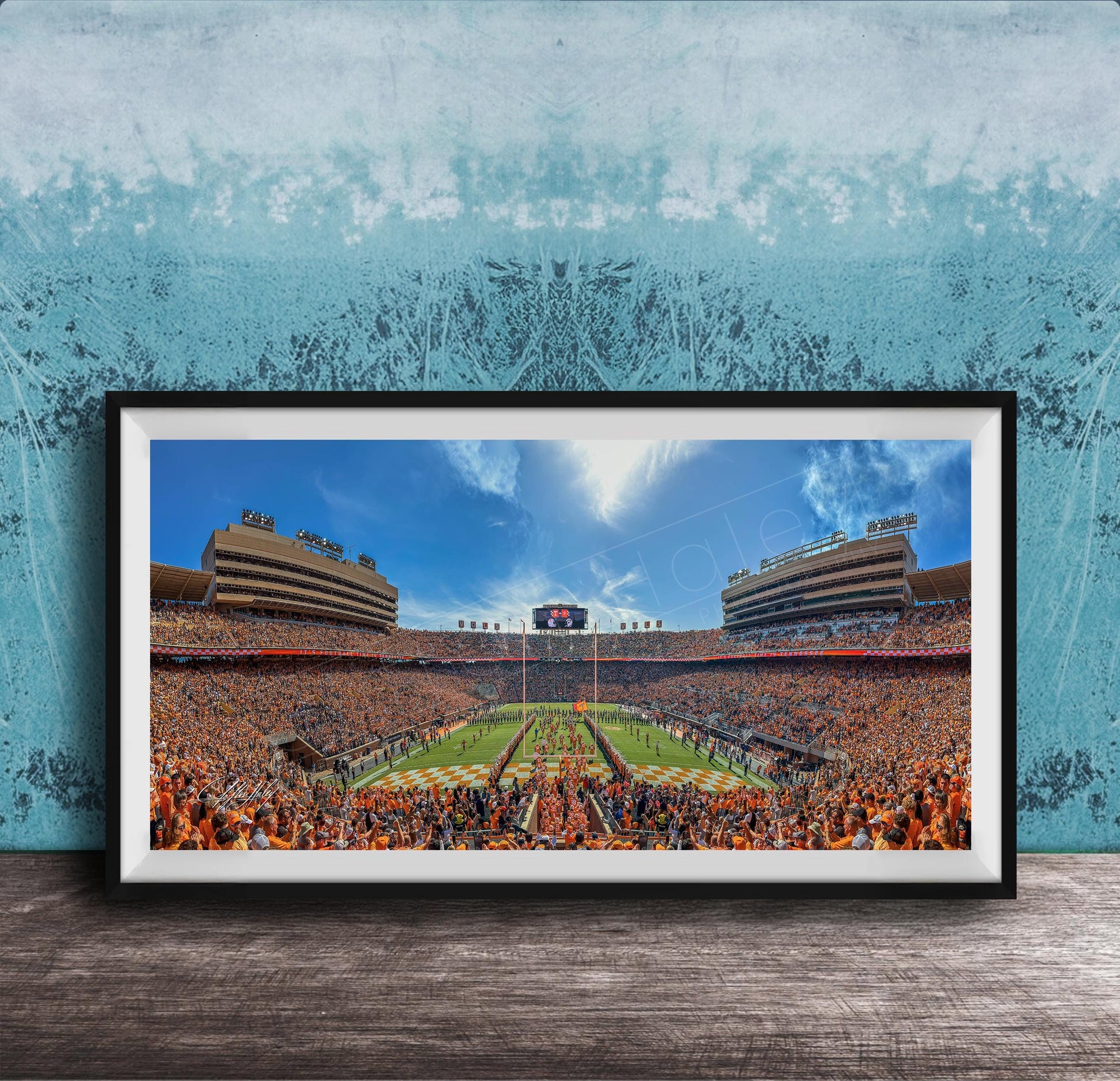 A framed photograph of a crowded football stadium with a large field and a scoreboard, set against a blue wall with a textured pattern.