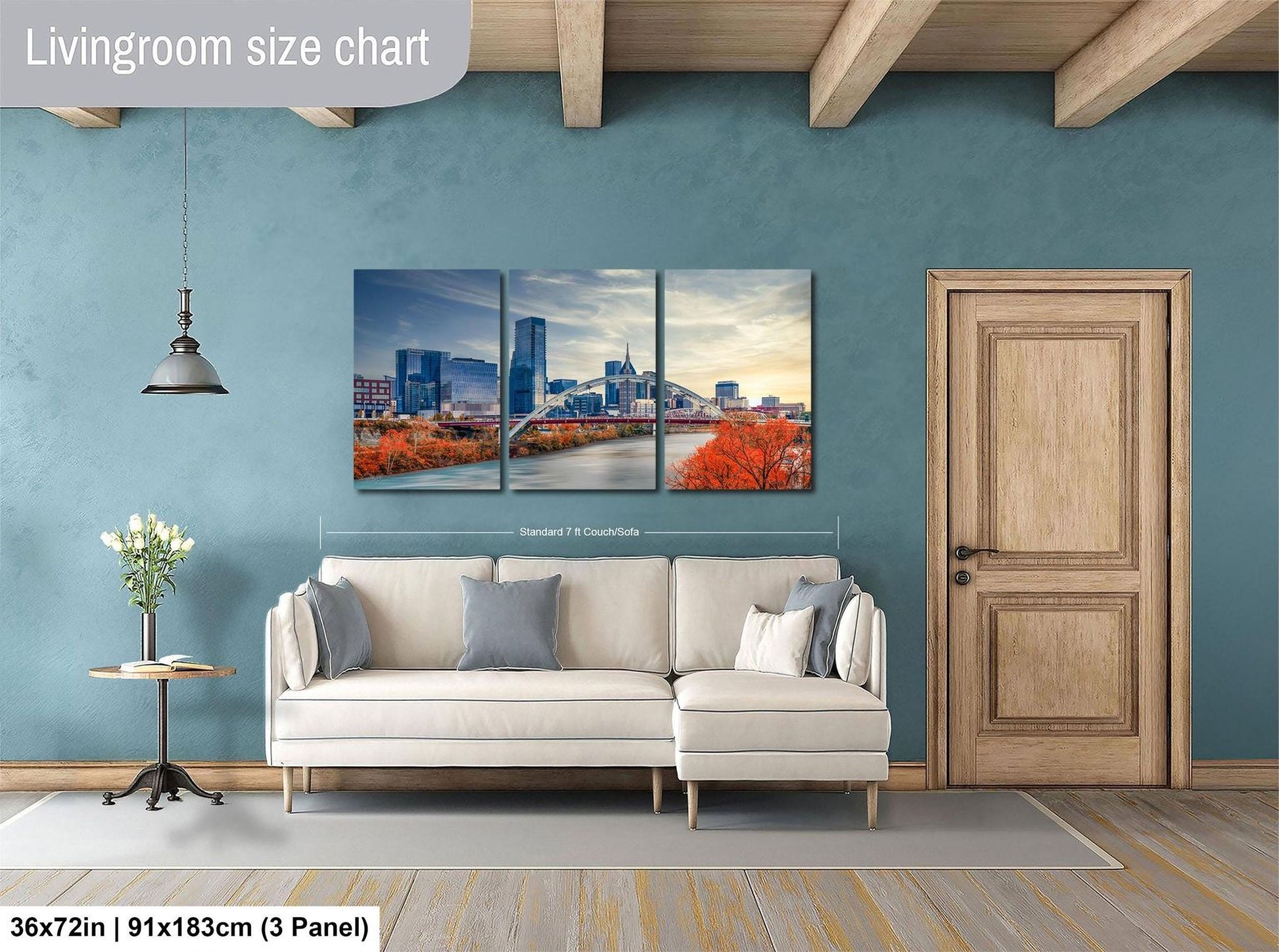 The image is a living room with a large wall art depicting a city skyline. The wall art is divided into three panels, each showing a different part of the cityscape. The room has a white sofa, a wooden door, and a small table with a vase of flowers. The wall art is approximately 36 inches wide and 11 inches tall, and it is mounted on a blue wall.
