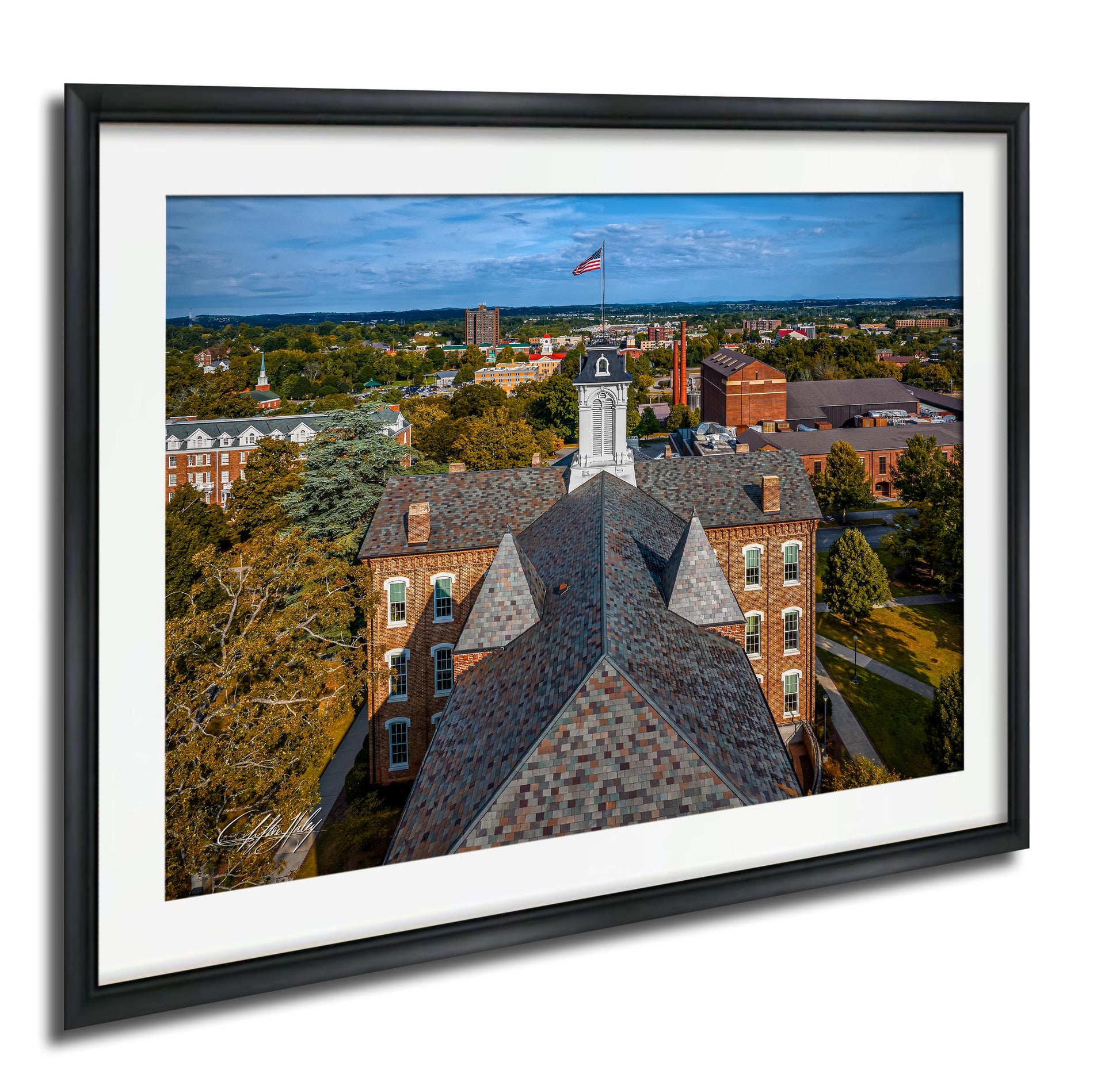 The image is a framed aerial view of a city, featuring a large brick building with a clock tower and a flag flying atop it. The surrounding buildings and greenery create a picturesque urban landscape.