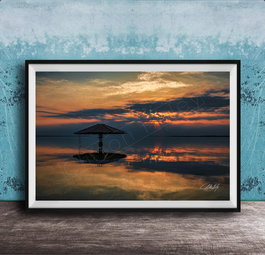 A framed photograph depicting a serene sunset over a calm body of water, with a lone umbrella standing in the foreground.