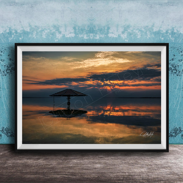A framed photograph depicting a serene sunset over a calm body of water, with a lone umbrella standing in the foreground.