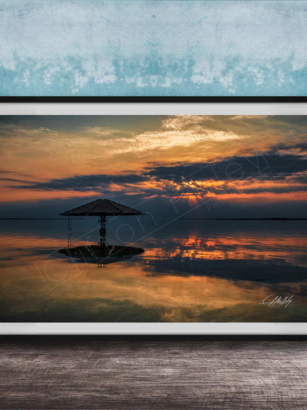 A framed photograph depicting a serene sunset over a calm body of water, with a lone umbrella standing in the foreground.