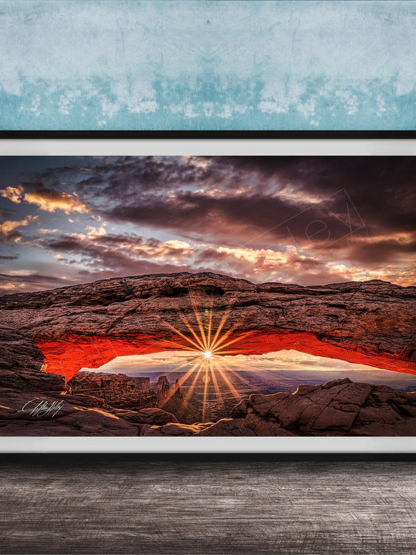 The image is a framed photograph depicting a dramatic sunset over a rocky landscape, with the sun's rays shining through the arch.