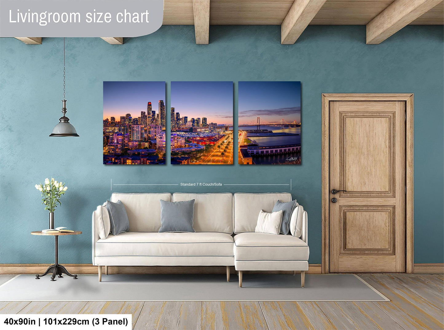 The image is a living room with a large wall art depicting a city skyline at night. The wall art is divided into three panels, each showing a different part of the cityscape. The room has a white sofa, a wooden door, and a wooden floor. The wall art is approximately 40 inches wide and 101.29 cm tall, with a 3-panel arrangement.
