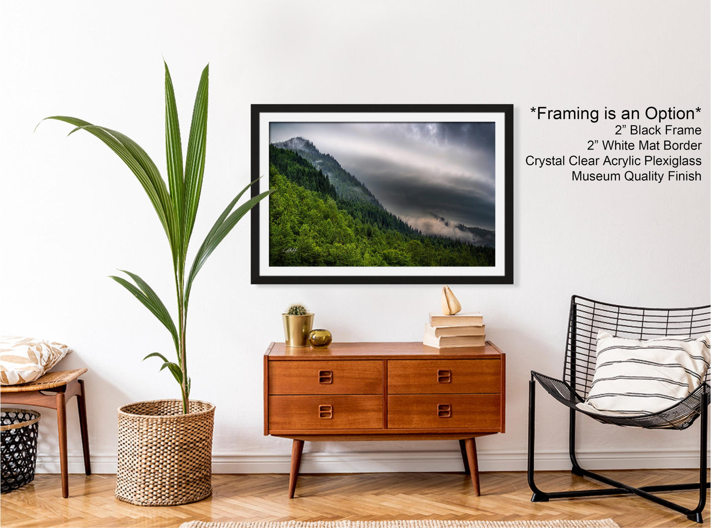 a framed photograph of a mountainous landscape with a cloudy sky, hanging on a wall above a wooden dresser with drawers. The dresser also has a wicker basket and a small plant. A black wire chair is positioned next to the dresser, and a stack of books is placed on top of the chair.