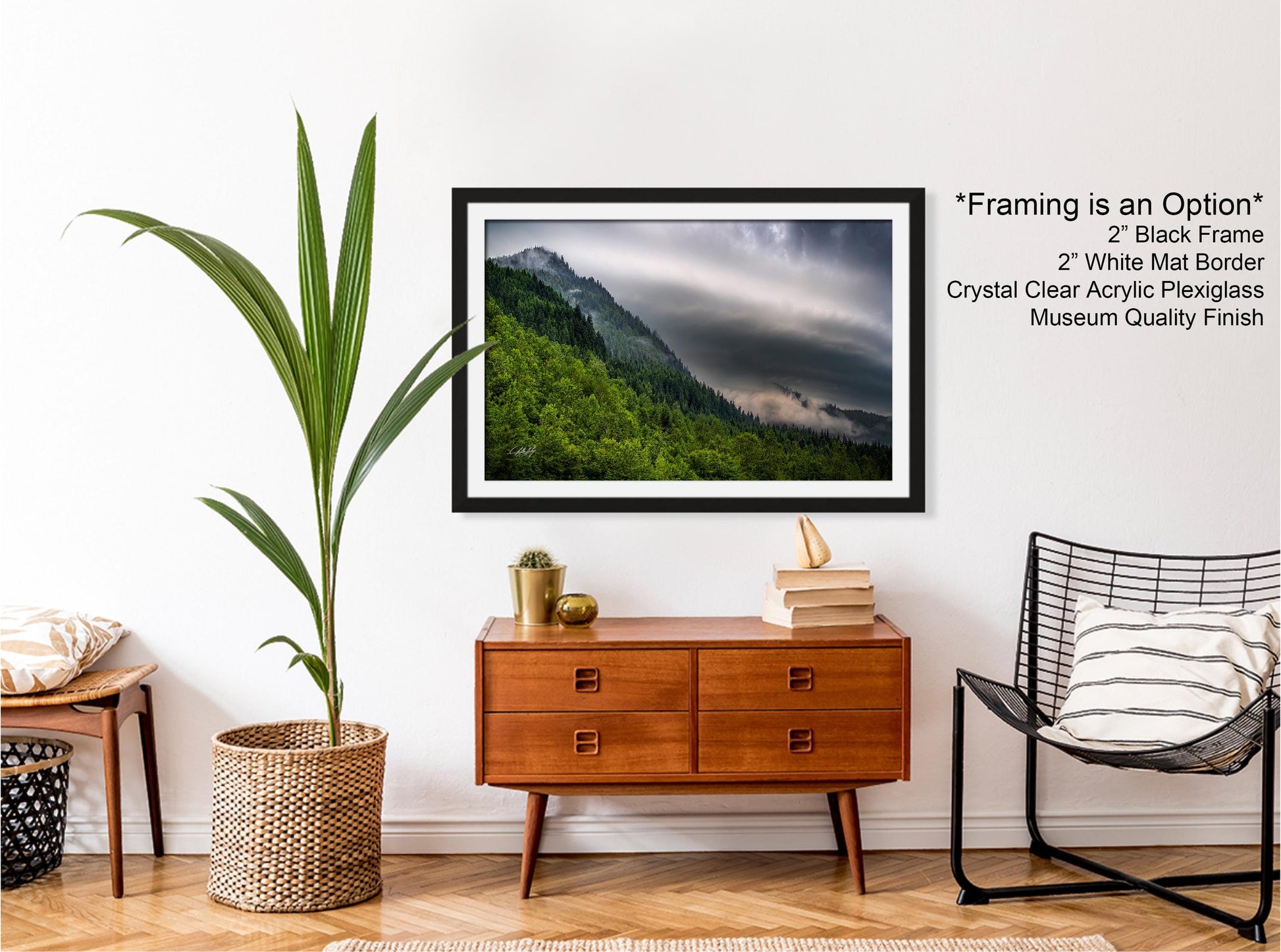 a framed photograph of a mountainous landscape with a cloudy sky, hanging on a wall above a wooden dresser with drawers. The dresser also has a wicker basket and a small plant. A black wire chair is positioned next to the dresser, and a stack of books is placed on top of the chair.