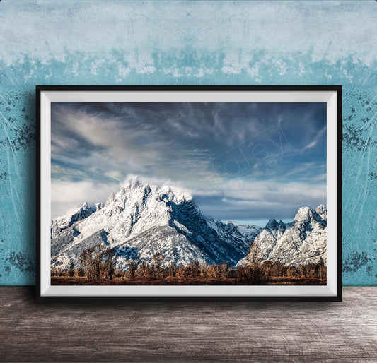 A framed photograph of a snow-capped mountain range against a cloudy sky, displayed on a wooden surface against a blue textured wall.