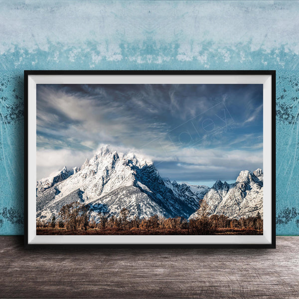 A framed photograph of a snow-capped mountain range against a cloudy sky, displayed on a wooden surface against a blue textured wall.