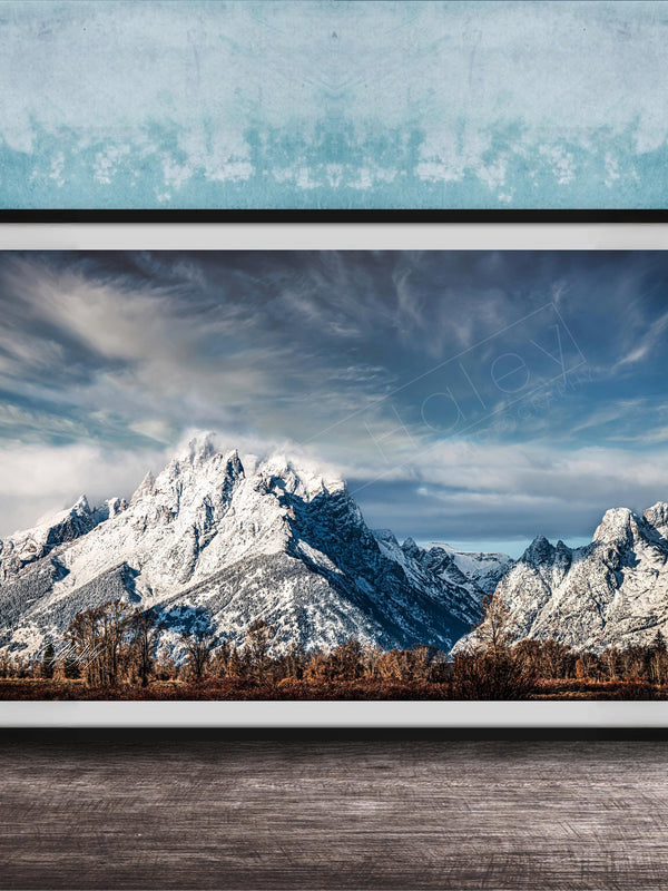 A framed photograph of a snow-capped mountain range against a cloudy sky, displayed on a wooden surface against a blue textured wall.