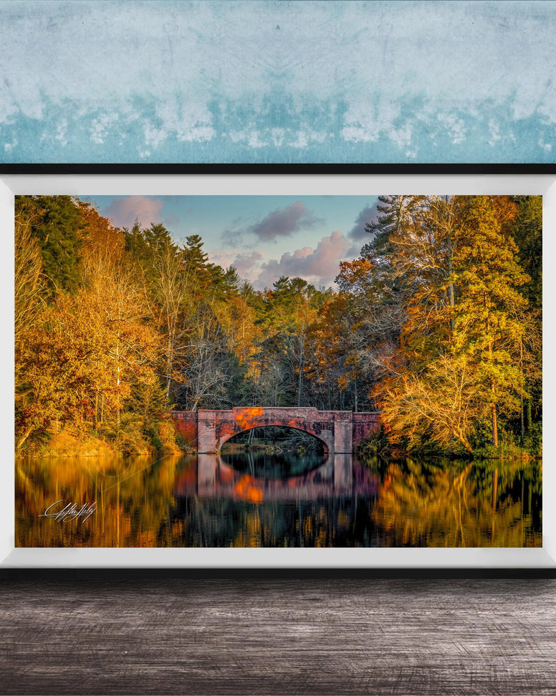 A framed photograph depicting a scenic autumn landscape with a red brick bridge spanning a tranquil lake, surrounded by vibrant fall foliage.