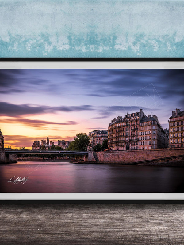 a picturesque cityscape at sunset, with a river running through the center and a row of buildings lining the riverbank. The sky is filled with warm, colorful hues, creating a serene and tranquil atmosphere. The buildings, with their unique architectural styles, add to the charm of the scene. The image is framed and displayed on a wooden surface, enhancing the overall aesthetic of the setting.