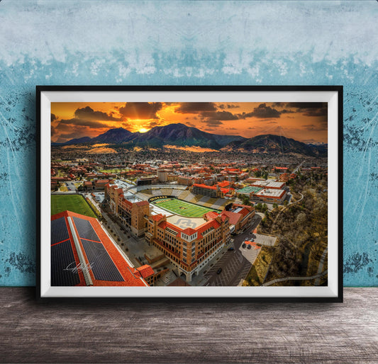 The image is a framed aerial view of a city with a large stadium, mountains in the background, and a dramatic sunset sky.