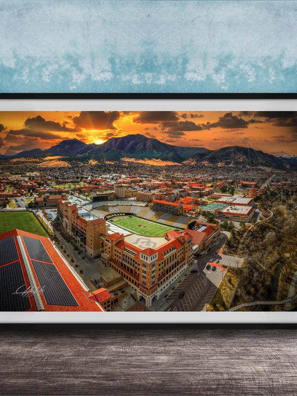 The image is a framed aerial view of a city with a large stadium, mountains in the background, and a dramatic sunset sky.