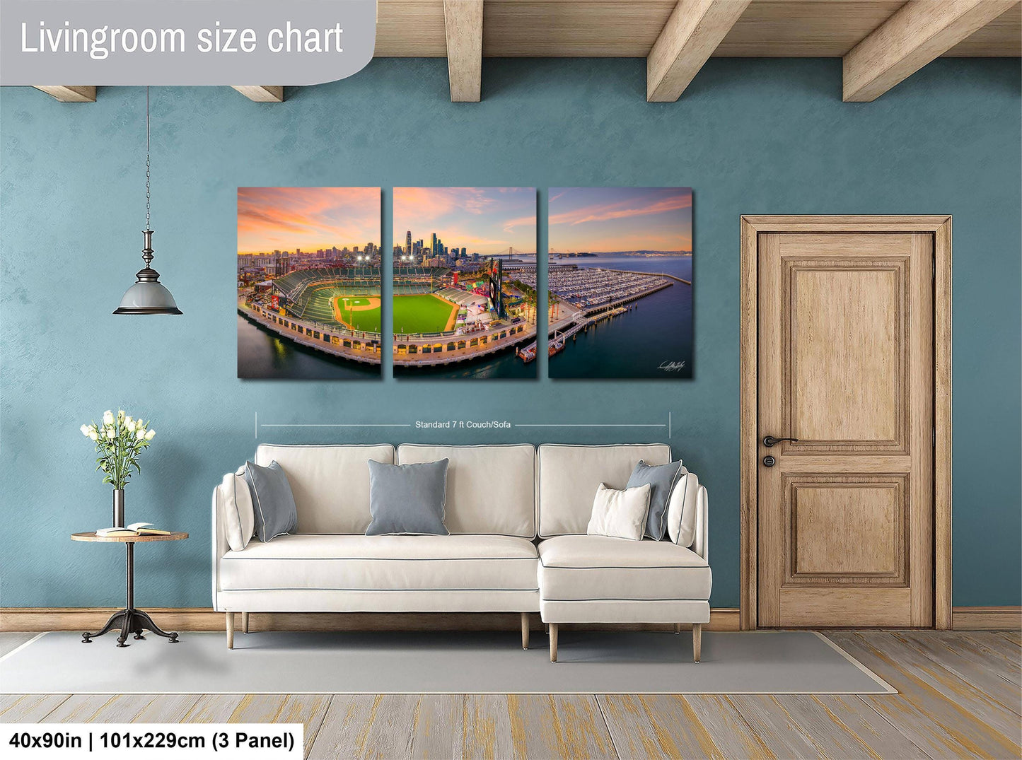 The image is a living room with a large, three-panel wall art depicting a baseball field and city skyline. The wall art is mounted above a white sofa, and there is a wooden door and a small table with a vase of flowers nearby. The wall art is approximately 40 inches wide and 101.29 cm tall, with a 3-panel layout.