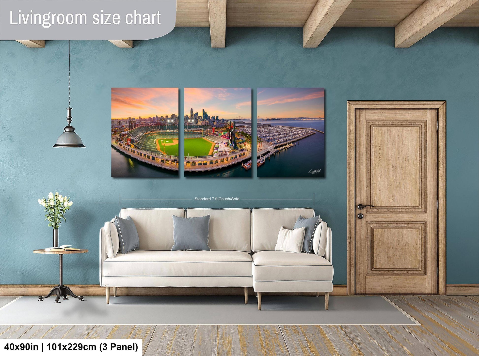 The image is a living room with a large, three-panel wall art depicting a baseball field and city skyline. The wall art is mounted above a white sofa, and there is a wooden door and a small table with a vase of flowers nearby. The wall art is approximately 40 inches wide and 101.29 cm tall, with a 3-panel layout.
