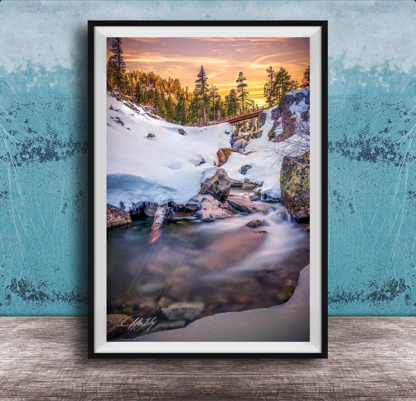 A framed photograph depicting a serene winter landscape with a snow-covered mountain, a flowing stream, and a vibrant sunset sky.