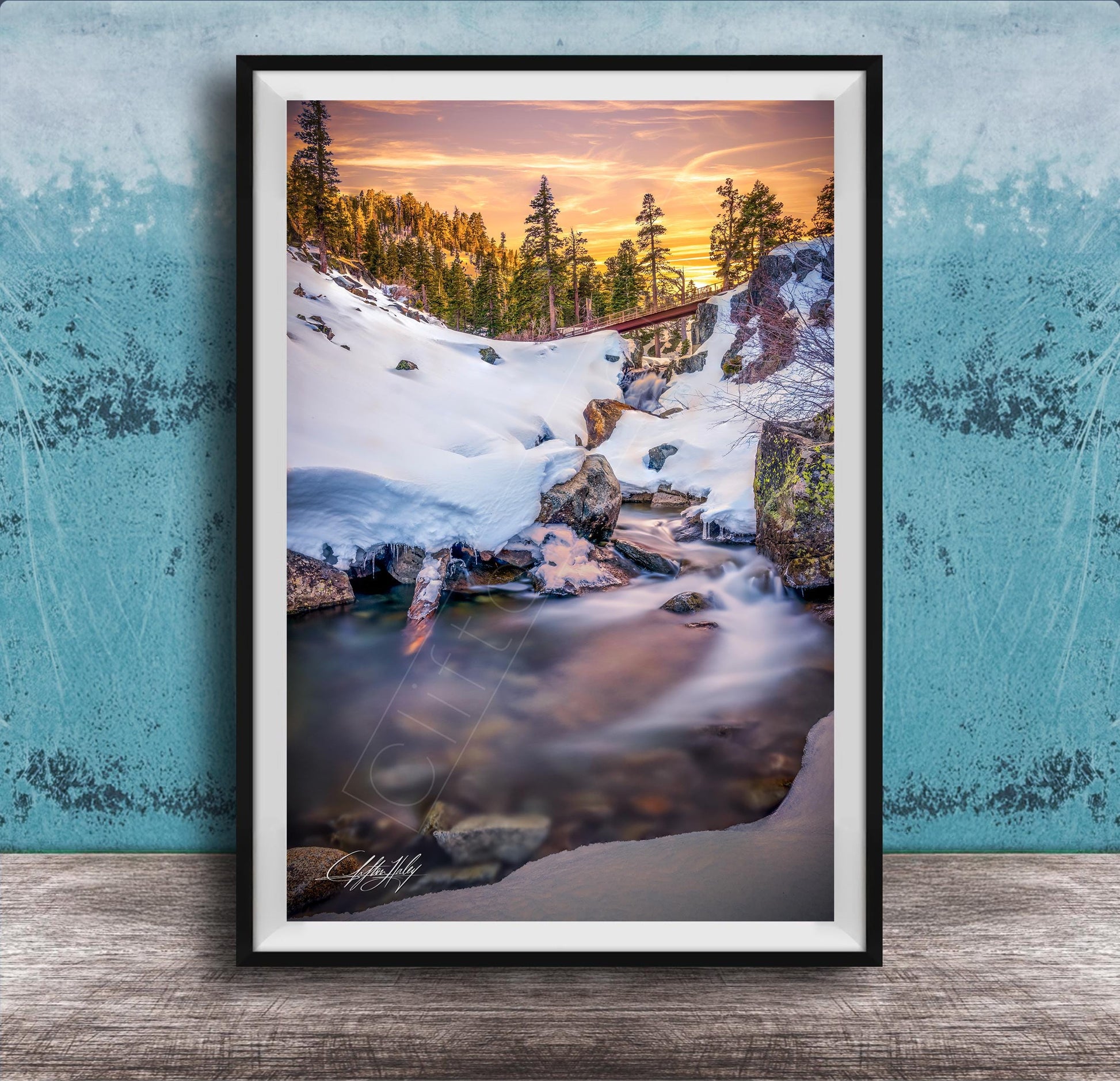 A framed photograph depicting a serene winter landscape with a snow-covered mountain, a flowing stream, and a vibrant sunset sky.