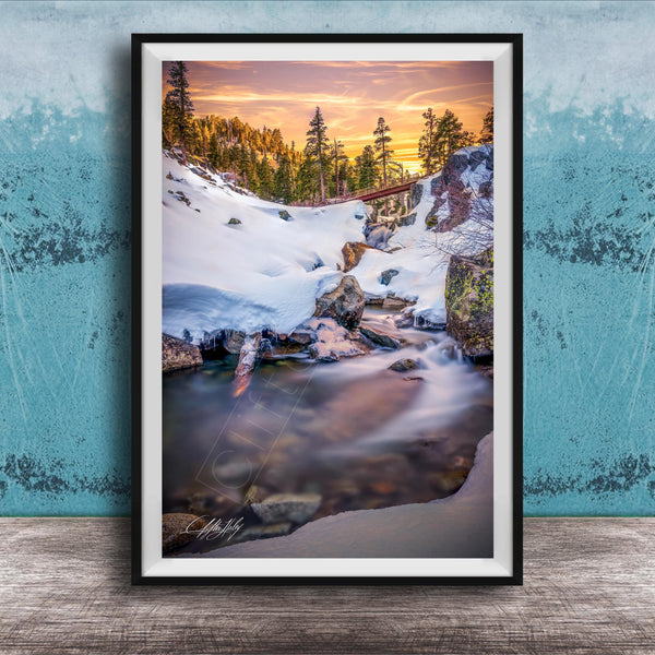 A framed photograph depicting a serene winter landscape with a snow-covered mountain, a flowing stream, and a vibrant sunset sky.