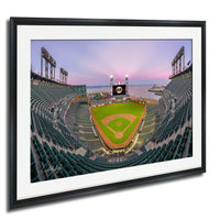 A framed photograph of a baseball stadium with a large scoreboard and field, set against a colorful sunset sky.