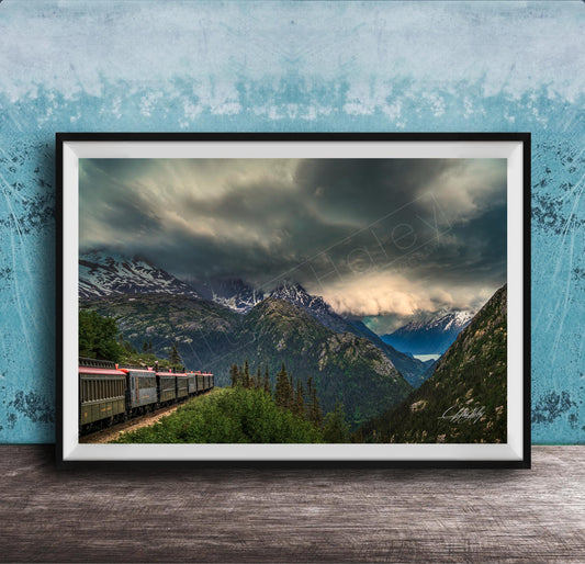 A framed photograph depicting a train traveling through a mountainous landscape with snow-capped peaks and a dramatic cloudy sky.