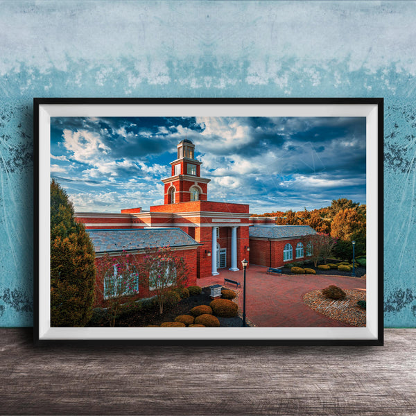 A framed photograph of a red brick building with a prominent tower, set against a blue sky with clouds.