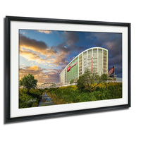 A framed photograph depicting a large building with a prominent red sign, set against a backdrop of a colorful sunset sky and lush greenery.