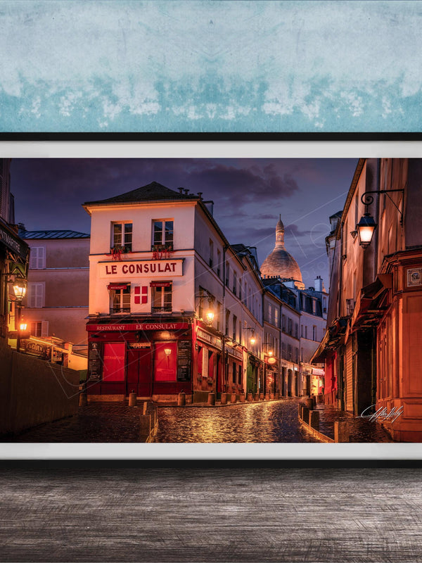 a charming street scene in Paris, France, with a row of colorful buildings lining the cobblestone street. The buildings are illuminated by warm street lamps, creating a cozy and inviting atmosphere. The sky above is a deep blue, providing a beautiful backdrop to the scene. The image is framed and displayed on a wooden surface, adding a rustic touch to the overall composition.