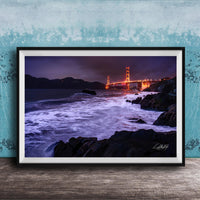 A framed photograph of the Golden Gate Bridge at night, with the bridge illuminated and its towers glowing against the dark sky. The waves are crashing against the rocky shoreline, creating a dramatic and serene scene.