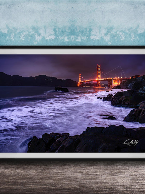 A framed photograph of the Golden Gate Bridge at night, with the bridge illuminated and its towers glowing against the dark sky. The waves are crashing against the rocky shoreline, creating a dramatic and serene scene.