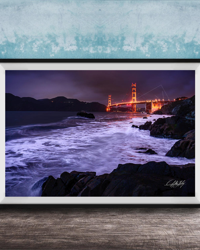 A framed photograph of the Golden Gate Bridge at night, with the bridge illuminated and its towers glowing against the dark sky. The waves are crashing against the rocky shoreline, creating a dramatic and serene scene.
