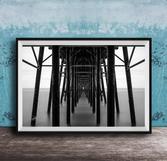 A black and white photograph of a long pier with multiple supporting columns, creating a sense of depth and perspective.
