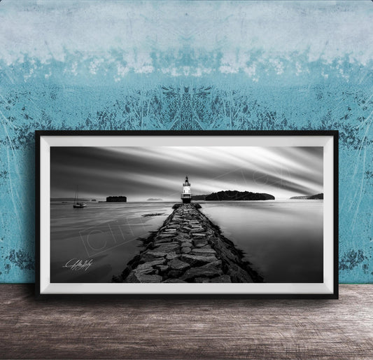 A black and white photograph of a lighthouse on a rocky pier, with a calm body of water and a cloudy sky in the background.